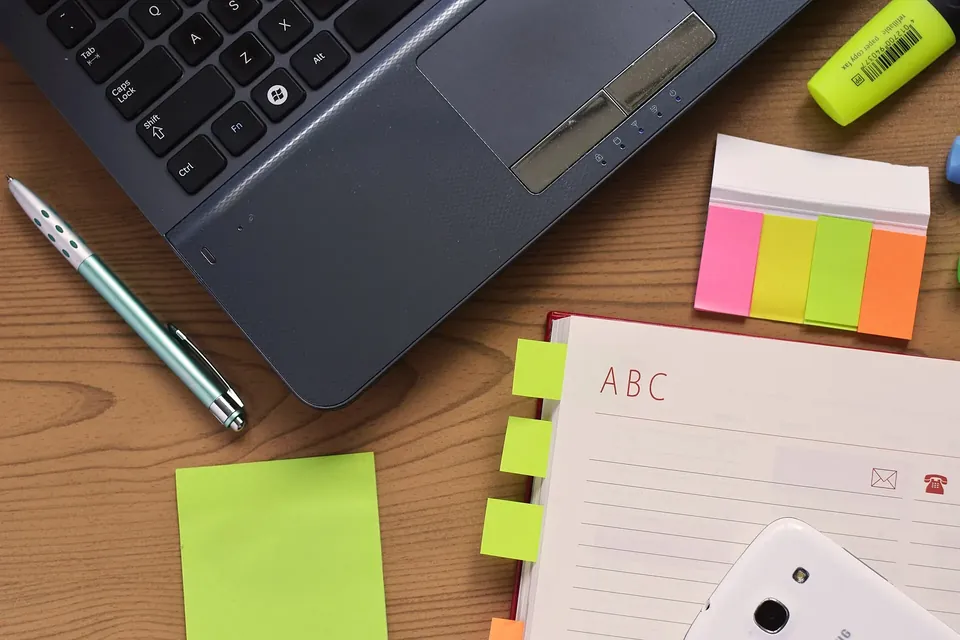 A laptop, a paper notebook, a pen and sticky notes on a wooden desk, seen from above